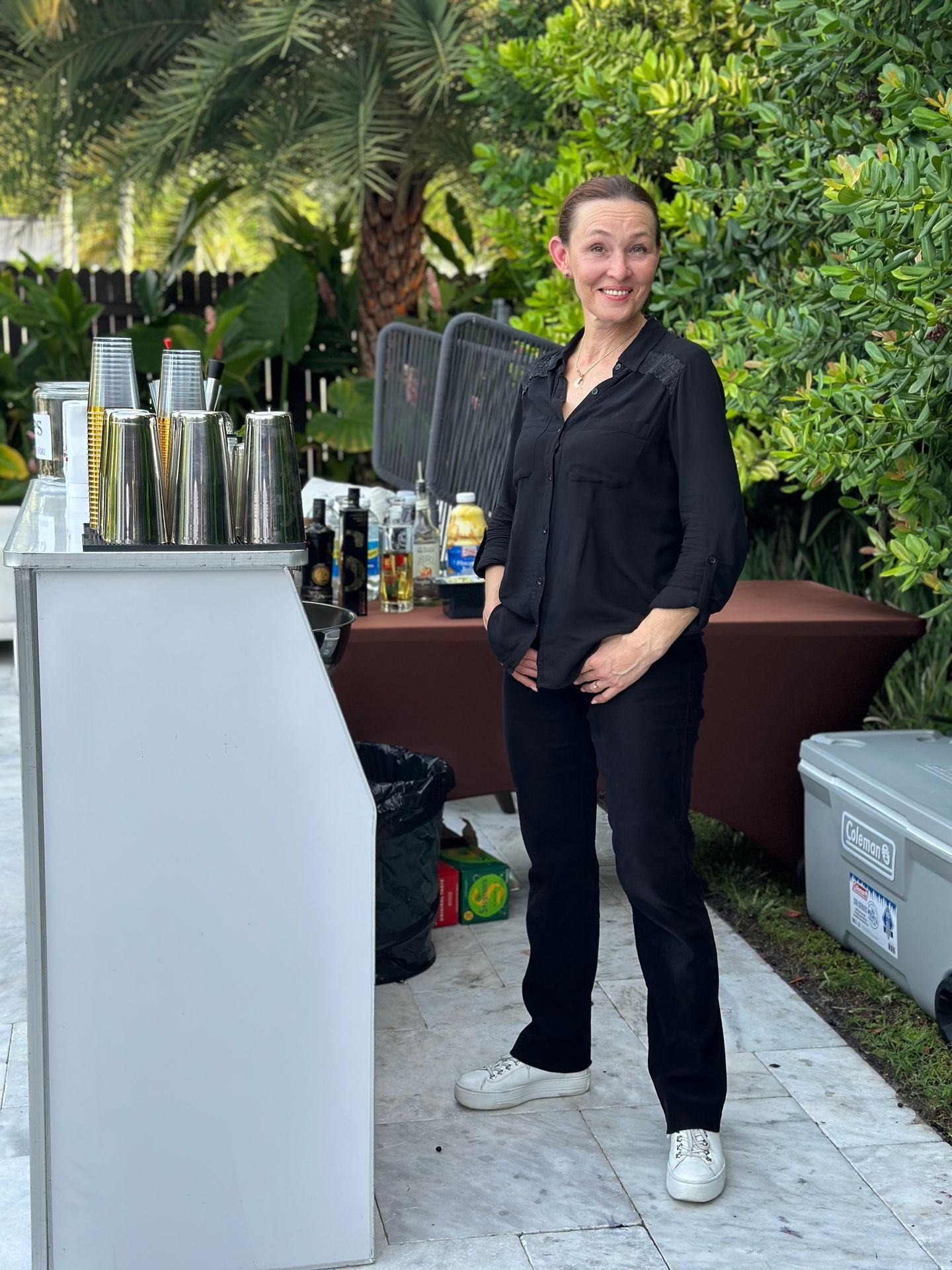 Smiling person in black outfit standing next to an outdoor bar setup with greenery in the background.