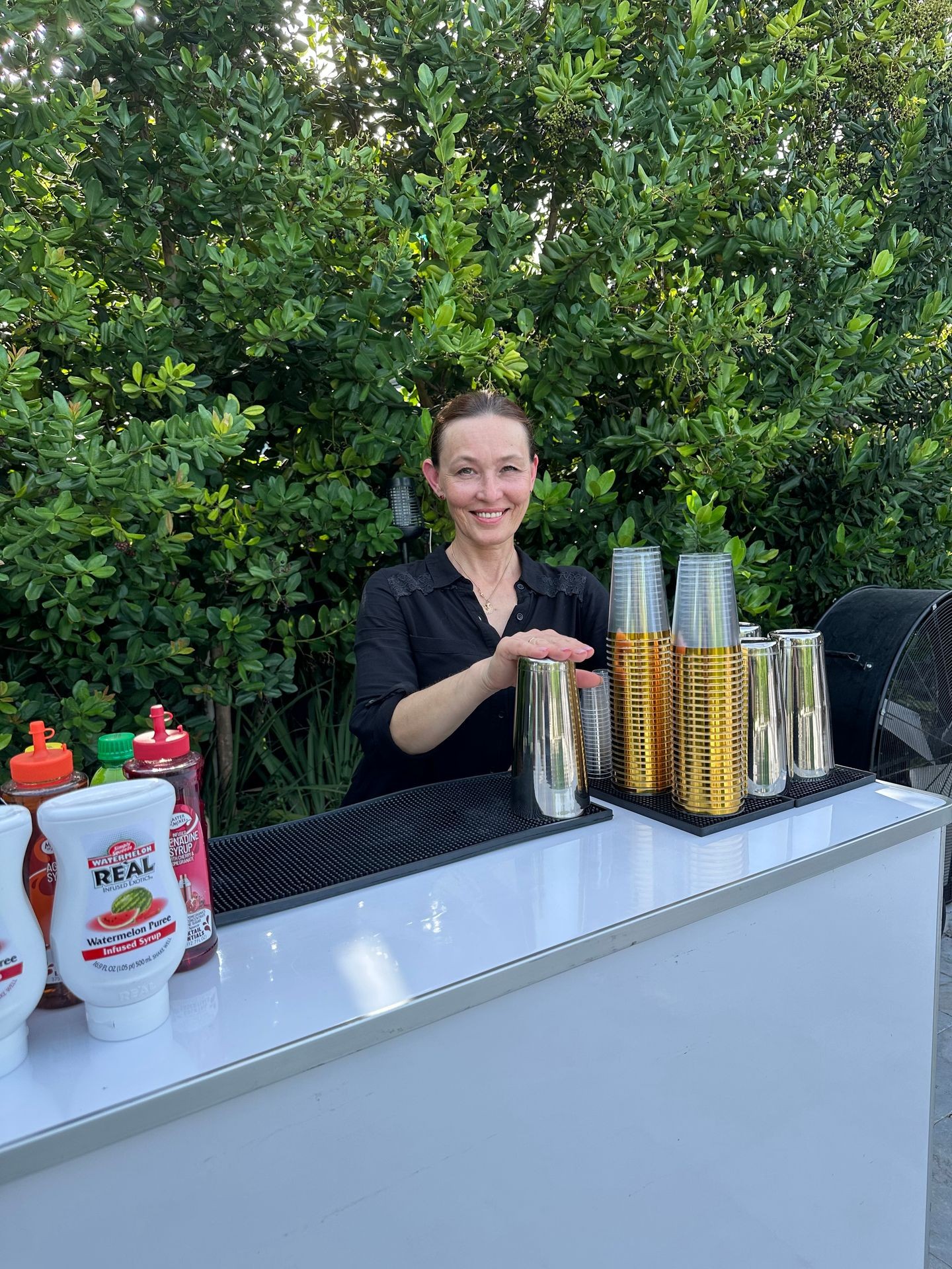 Woman behind outdoor bar with stacked cups and drink bottles, surrounded by lush greenery.