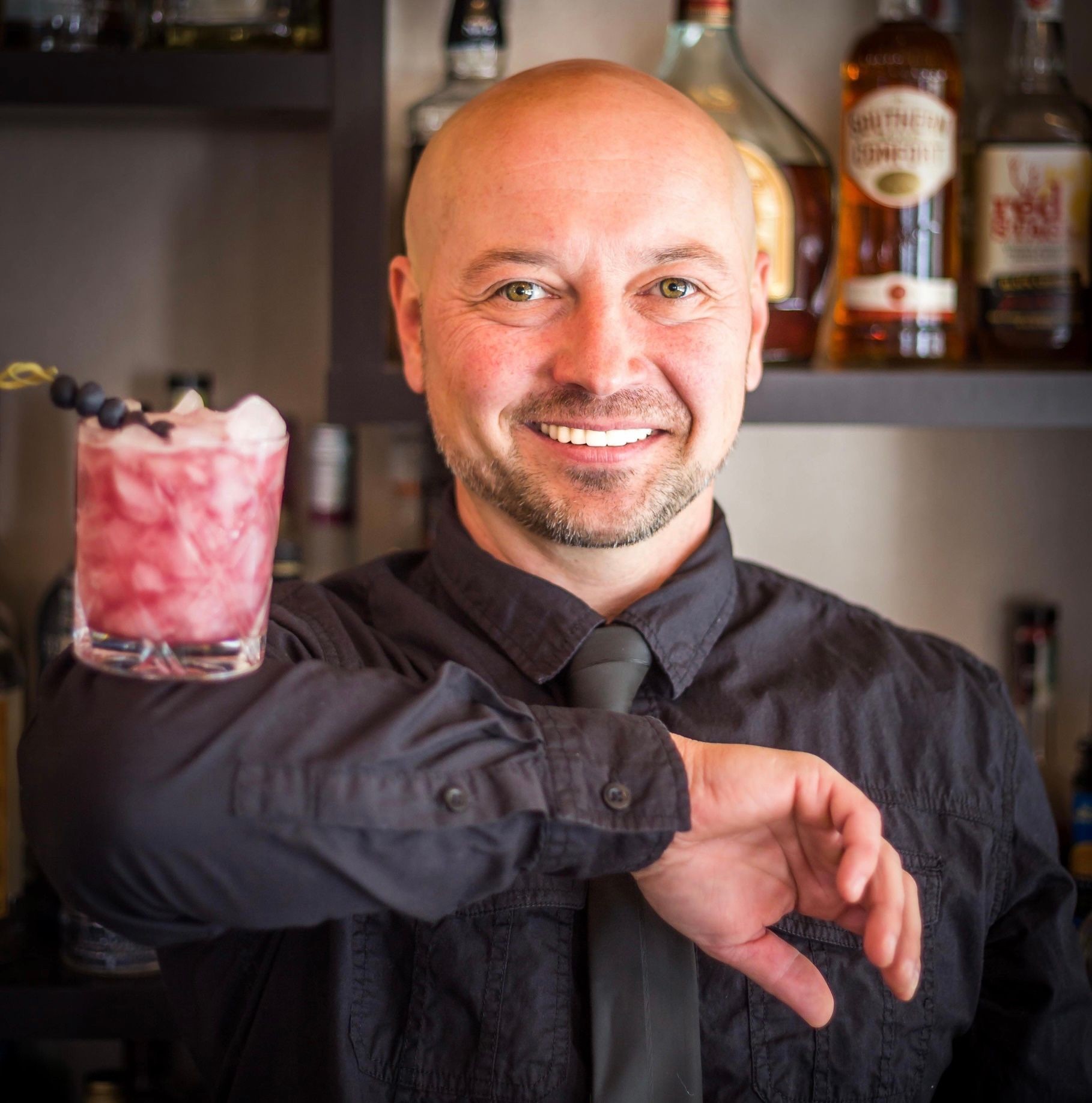 Man balancing a cocktail on his forearm, smiling, with bottles on shelves in the background.