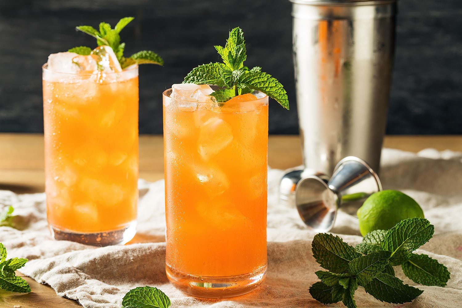 Two orange cocktails with ice and mint garnish, beside a shaker and lime on a cloth.