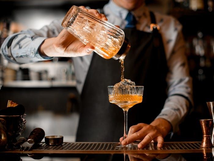 Bartender pouring a cocktail from a shaker into a glass with ice at a bar.