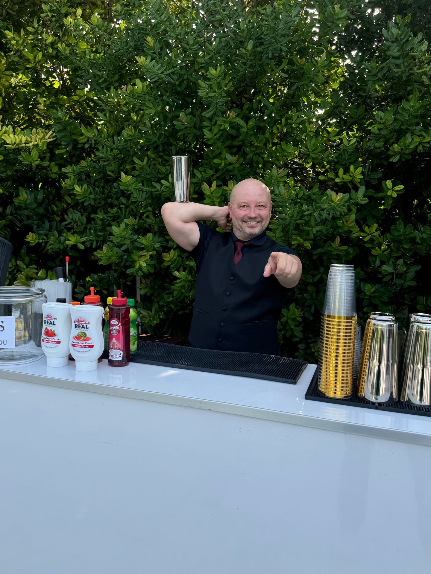 Bartender performing a flair bartending trick with a cocktail shaker behind an outdoor bar setup.