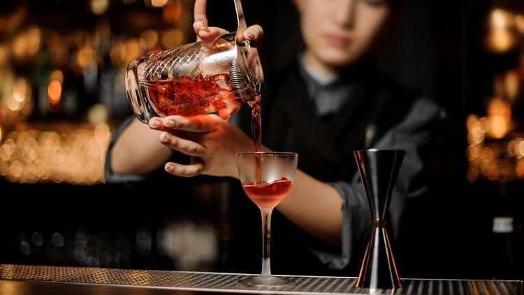 Bartender pouring a red cocktail into a glass at a bar with dim lighting.