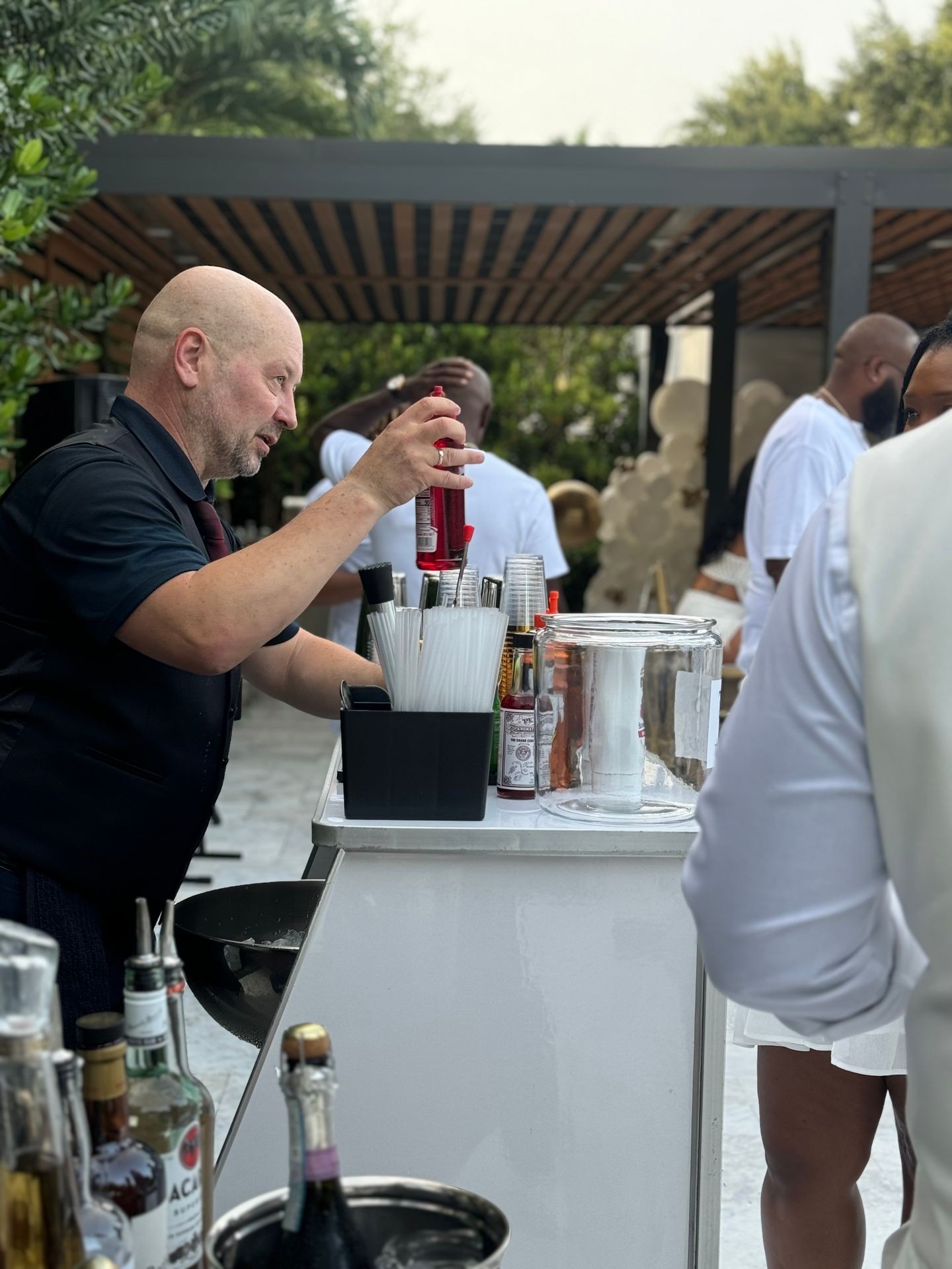 Bartender preparing drinks at an outdoor event with a variety of beverages and greenery in the background.