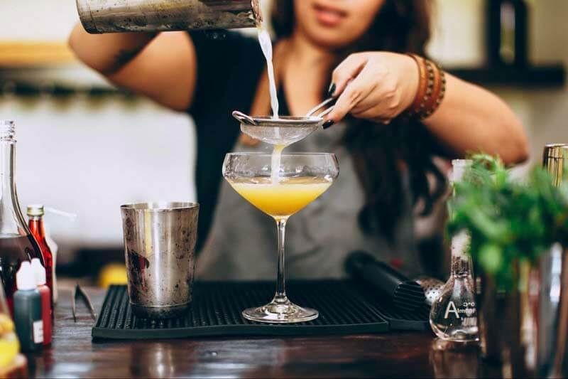 Bartender straining and pouring a cocktail into a glass on a bar counter.