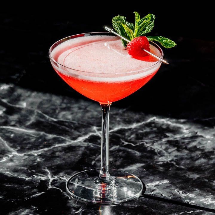 Elegant pink cocktail in a glass garnished with raspberry and mint on a dark marble surface.